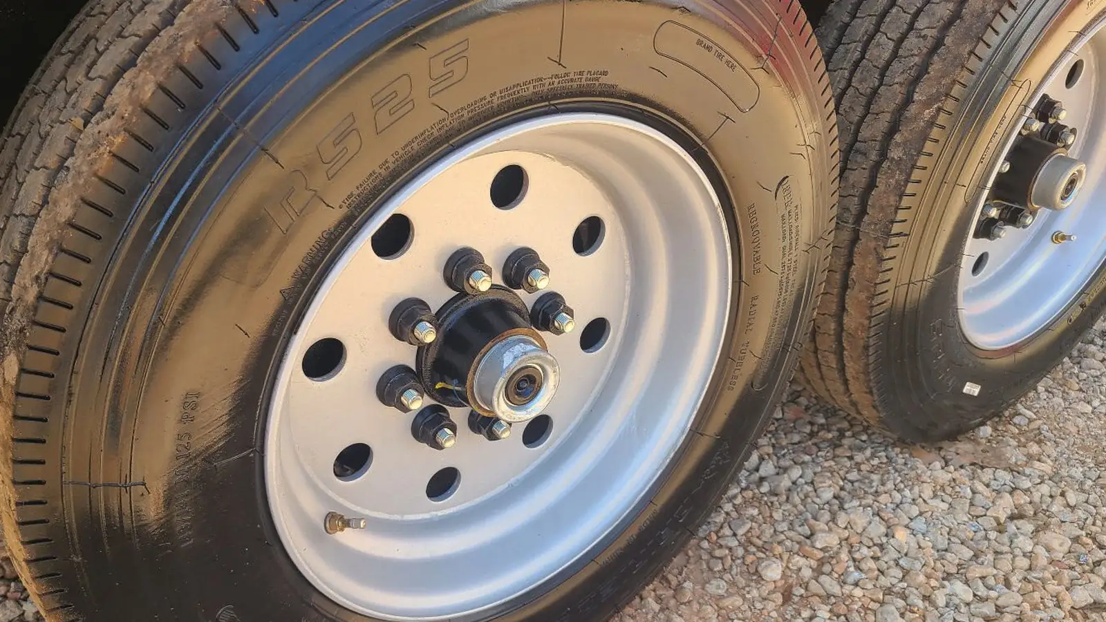Close view of an eight lug hub on a silver modular steel wheel of a tandem axle dump trailer, with eight studs and lug nuts spaced evenly around the black hub face, the grease cap in the center, and the second wheel of the tandem behind it