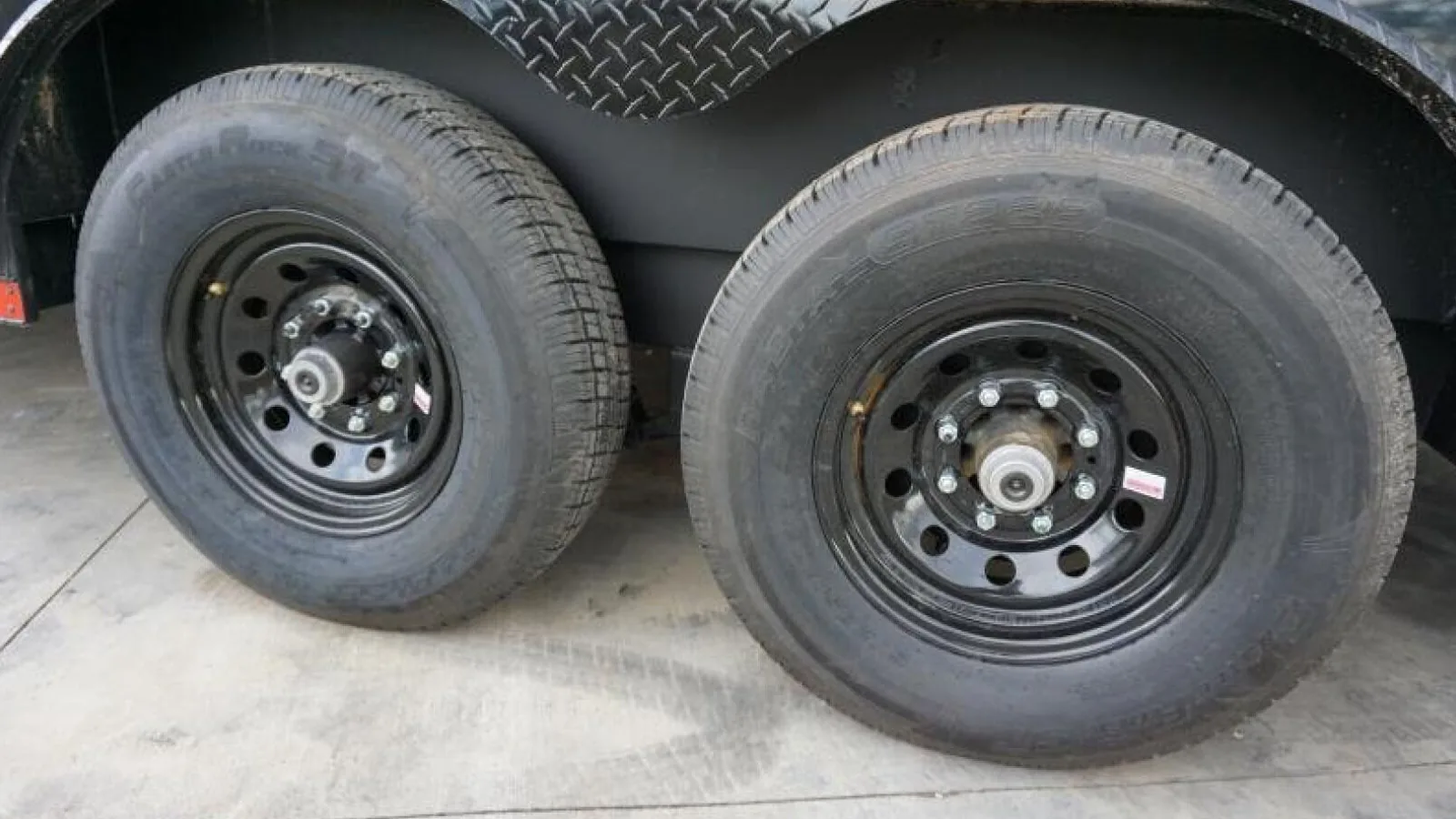 The tandem axle hubs and wheels under a black enclosed cargo trailer on the lot at Trailer Source in Greenville, South Carolina, with the grease caps and lug nuts in clear view