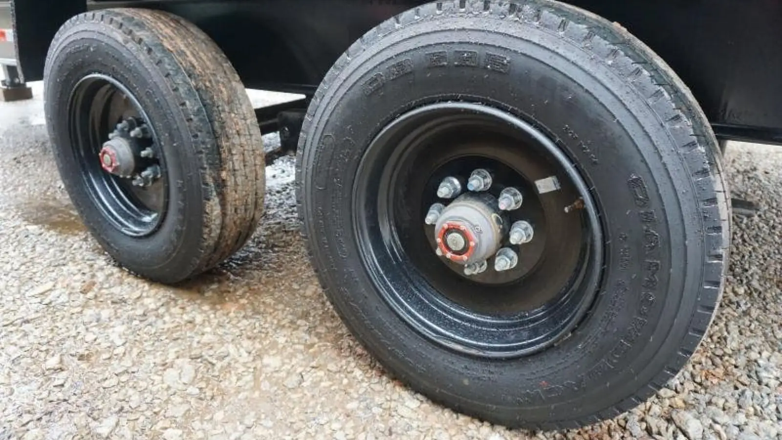 Low view of the near side of a tandem axle under a black dump trailer on a wet gravel lot, showing both wheels, a hub with a red bearing protector cap at the center of each one, and part of the axle tube in the gap between the tires